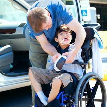 Man assisting a smiling boy in a wheelchair outside near a car.