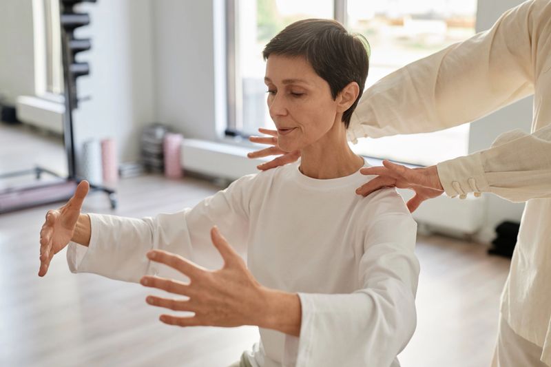 Waist up shot of brunette senior woman doing qigong concentration exercise and holding hands in air while unrecognizable trainer adjusting position of her shoulders