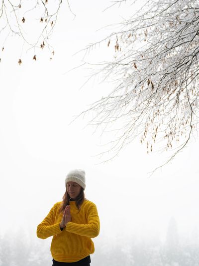 Woman in yellow sweater meditating in snowy forest with eyes closed.