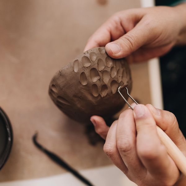 Hands sculpting textured patterns on a clay bowl using a carving tool.