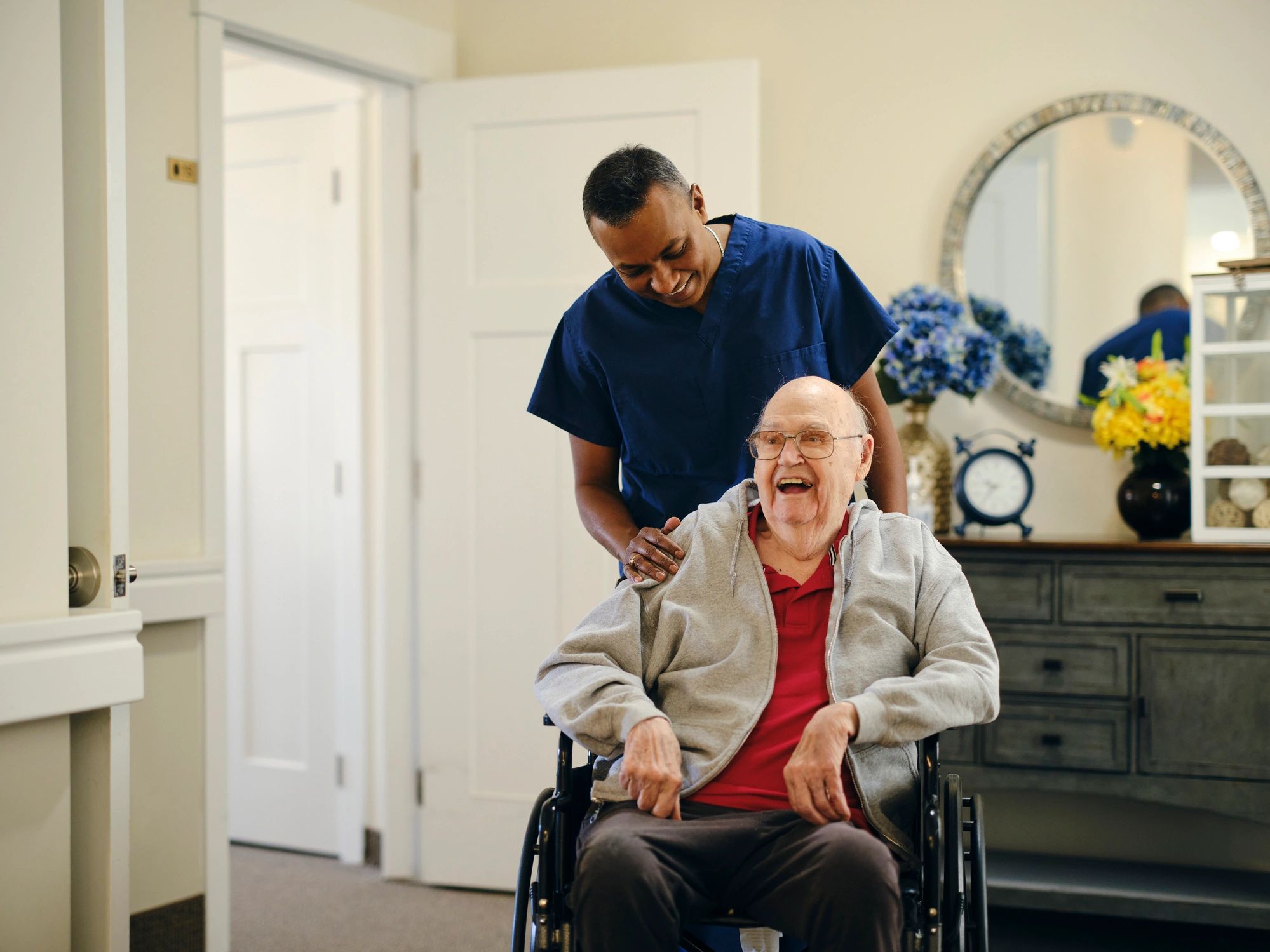 Caregiver smiling while pushing an elderly man in a wheelchair.