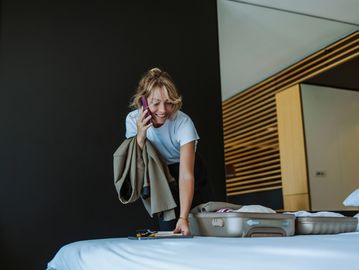 Smiling woman talking on phone while packing a suitcase on a bed.