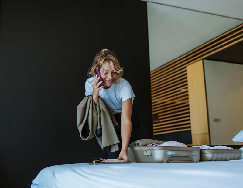 Close up shot of a beautiful blonde woman standing in the hotel room talking on the phone. She is packing a suitcase that is on the bed and holding a coat in another hand. She is looking down and smiling.
