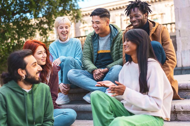 A diverse group of friends laugh and converse on sunny city steps, enjoying a lively discussion.