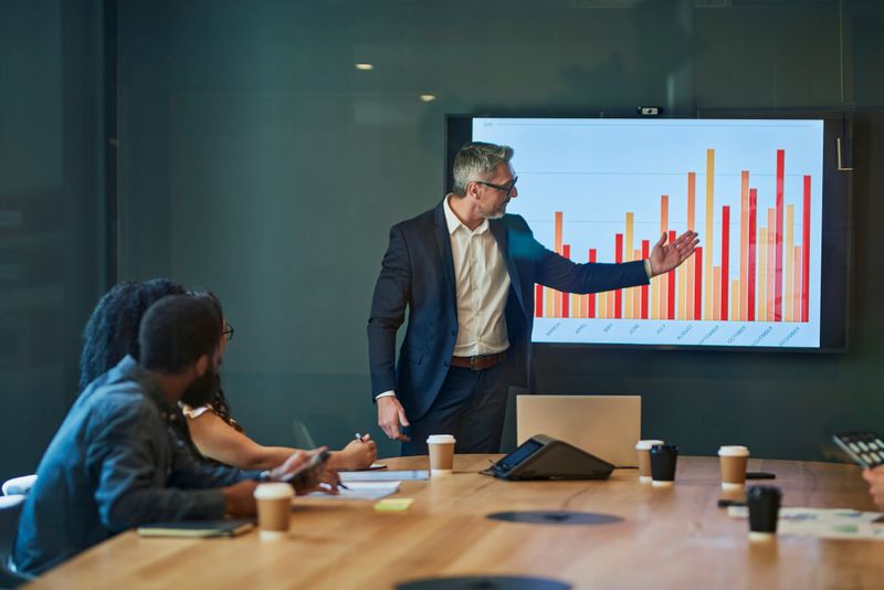 Confident businessman leading a meeting and presenting financial data to colleagues in a modern office. The group is seated around a conference table and looking at a large screen displaying graphs and charts.