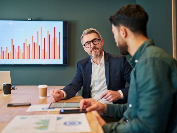Two men discussing business data in a meeting room with charts displayed.