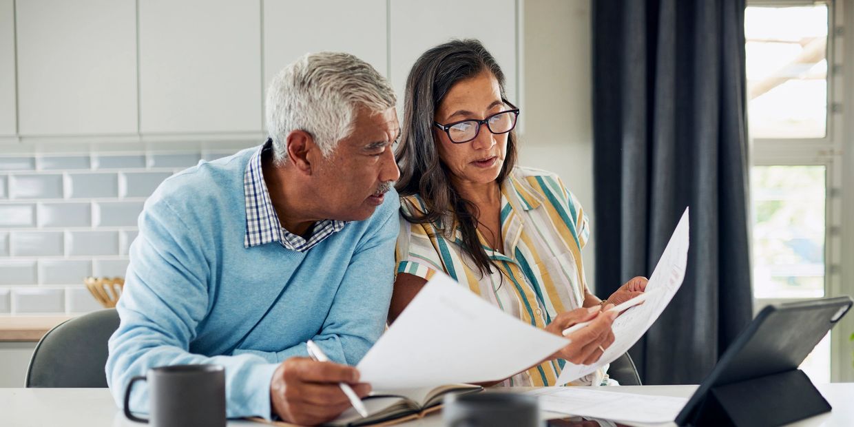 An elderly couple reviews documents together at a kitchen table with a tablet and coffee mugs.