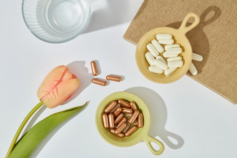 The hard capsule is placed in a blue plate, the white pill is placed in a yellow plate, a glass of water and a tulip branch on a white background. View from above.
