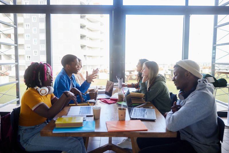 Diverse group of young college students talking while studying together around a table in a school cafeteria