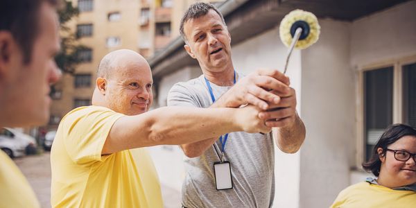 Man helps another man use a cleaning tool outdoors, both smiling.