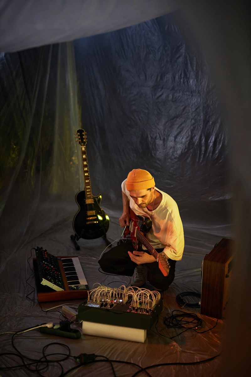 Vertical high angle shot of adult male musician with electric guitar setting up equipment for sound check in dim light makeshift studio with plastic cover