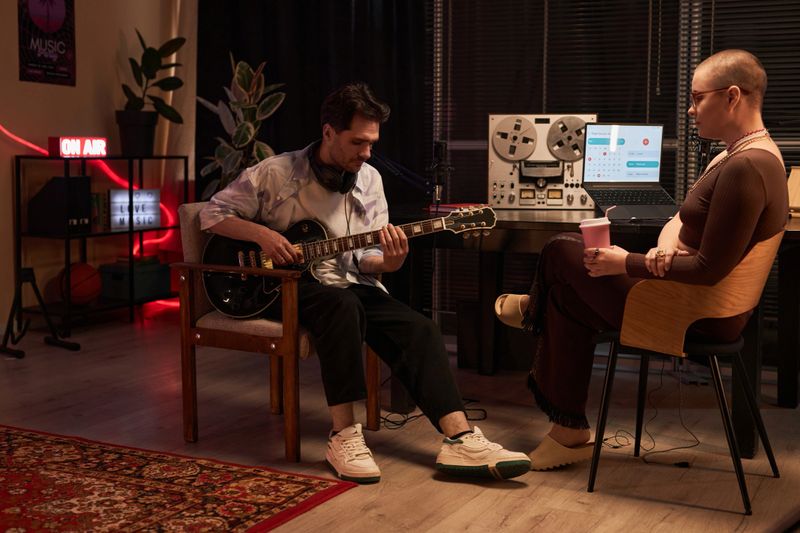 Wide shot of adult male composer with headphones on neck playing semi-acoustic guitar while sitting with female interviewer in podcast room
