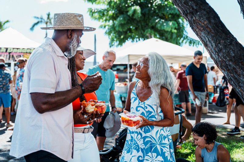 An active and adventurous African American senior couple eat fresh fruit and cheerfully chat with their local tour guide, a senior woman of Hawaiian decent, while at a farmer's market in Honolulu, Hawaii.