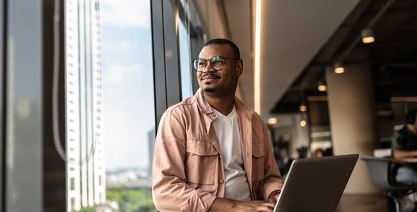 Man with glasses working on a laptop by a large window in a modern office.