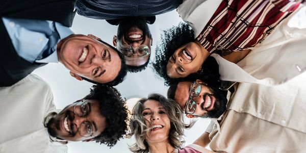 A diverse group of six people smiling in a circle, looking down at the camera.
