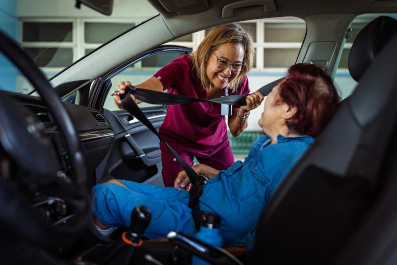 A kind occupational therapist of Hawaiian and Finnish descent smiles as she helps a female geriatric patient get seated and buckled into the passenger seat of a car.