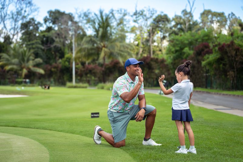 A kindergarten aged girl of Asian ethnicity stands beside her proud, loving father, who is kneeling on the golf course, high-fiving each other over her accomplishments.