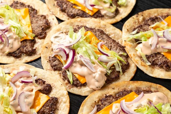 Burger tacos with ground beef, cheese, lettuce, red onion, and sauce on tortillas.