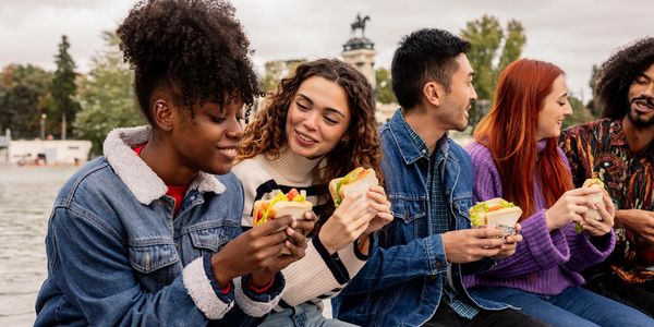 A group of friends enjoying sandwiches together outdoors by the water.