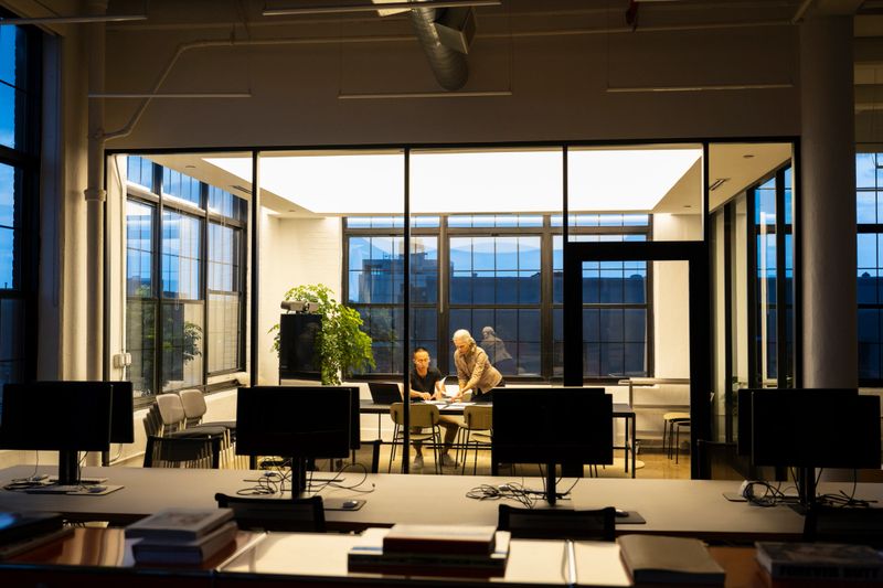 View across dark office as mature marketing director and young assistant manage paperwork for project development.