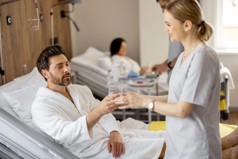 Nurse gives a drink for a male patient lying in comfortable bed in medical ward. Concept of medical support and assistance during the rehabilitation period after surgery