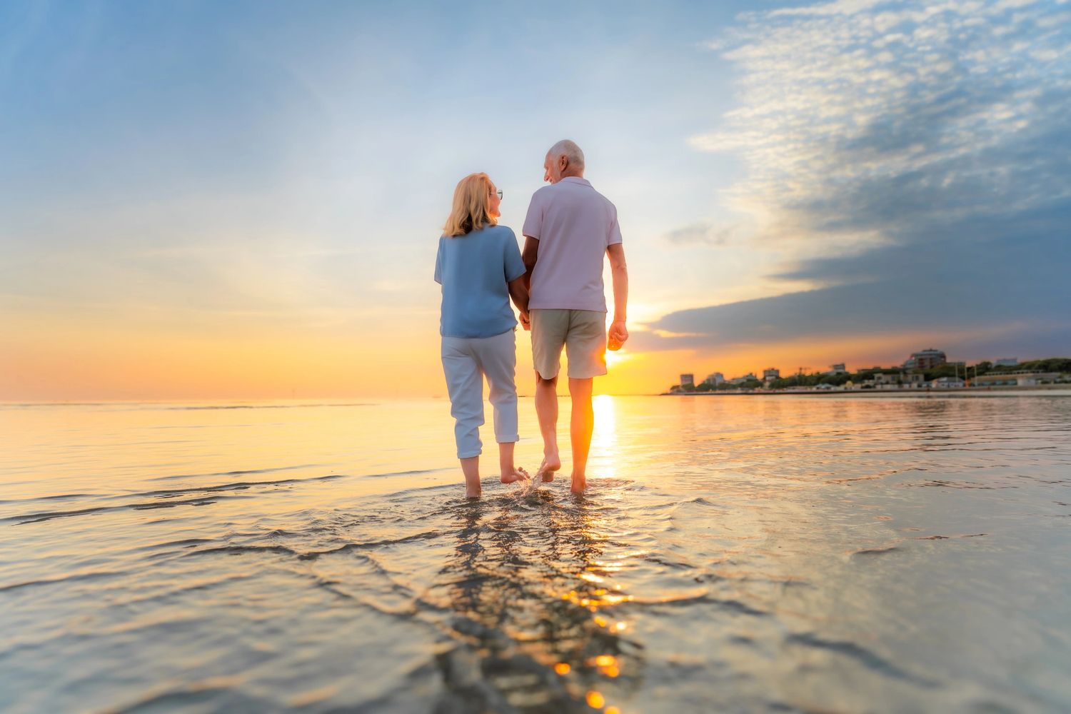 Elderly couple walking hand-in-hand in shallow water at sunset.