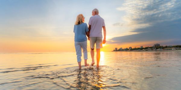 Elderly couple walking barefoot on beach at sunset.