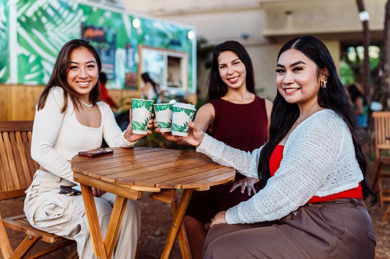 Three young women of multiracial ethnicity cheers their beverages, sitting at an outdoor table and smiling directly at the camera.