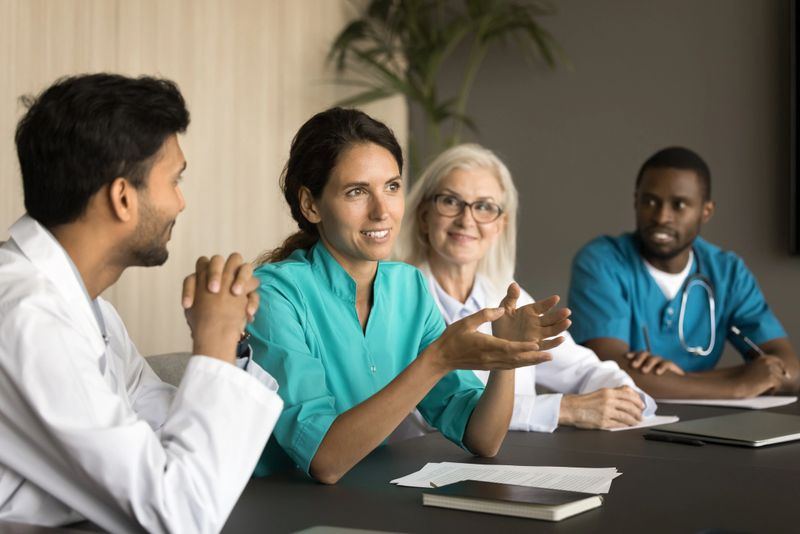 Positive young Latin surgeon woman speaking at professional meeting to multiethnic colleagues, offering ideas for collaboration, brainstorming. Diverse practitioners discussing doctor occupation