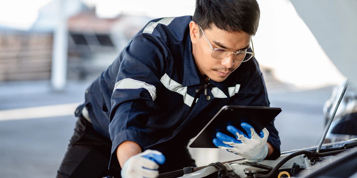 A mechanic wearing gloves and glasses uses a tablet while inspecting a car engine under the hood.