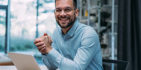 Smiling man with glasses sitting at a desk with a laptop in a bright office.