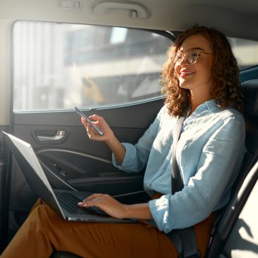 Woman working on laptop and phone inside a car, smiling.