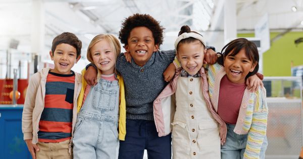 Five diverse kids smiling brightly with arms around each other indoors.