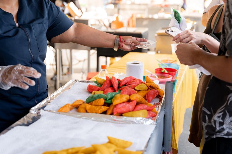 At a street stall, a Latin empanadas vendor hands change to a customer after a transaction