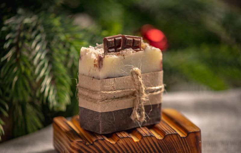 Natural handmade chocolate soap on an organic wooden soap dish against the background of green branches of a coniferous tree.