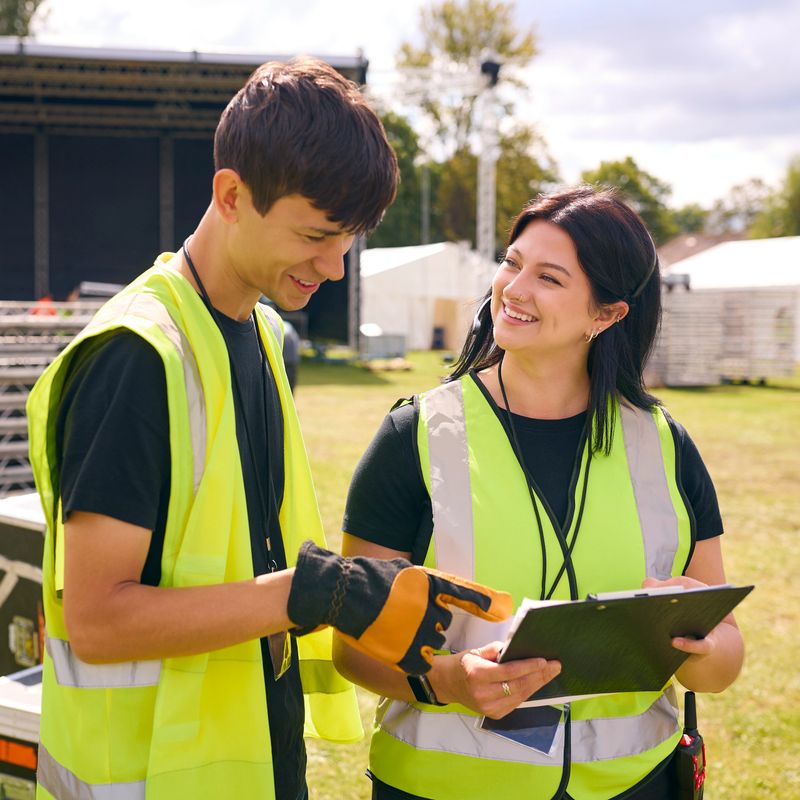 Male And Female Production Team With Headsets Setting Up Outdoor Stage For Music Festival Or Concert