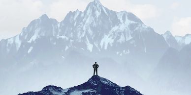 A man stands on a mountain, eyeing a distant summit with a red flag.