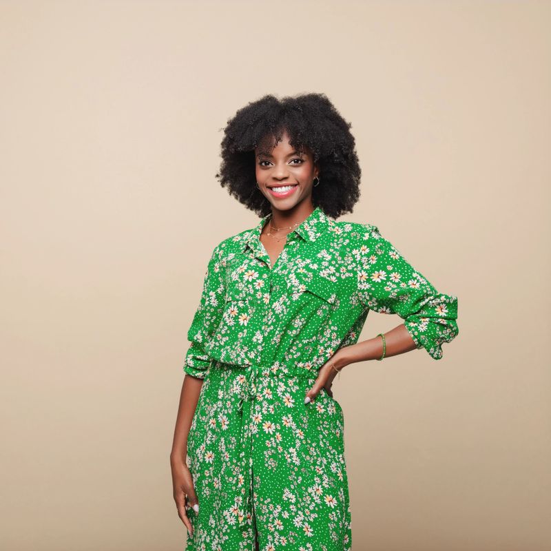 Beautiful black young woman wearing green dress standing against beige background with hand on hip and smiling at camera.