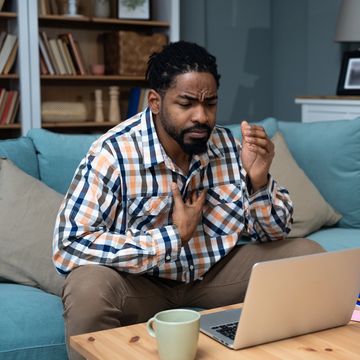 Man clutching his chest, looking distressed on a couch with a laptop.