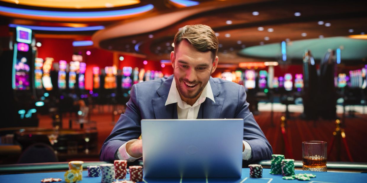 Man in suit smiling at laptop in casino with poker chips.