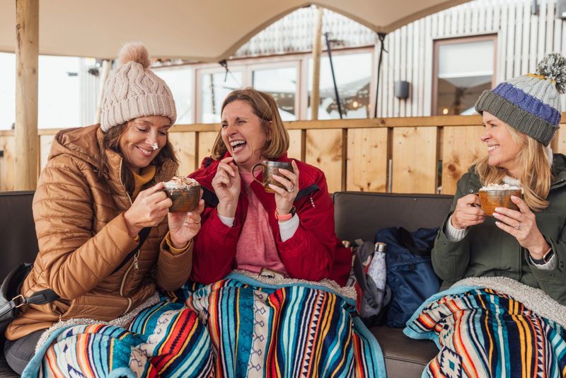 Three women sitting at a cafe drinking hot chocolate with whipped cream on the top. One has cream on her nose and the other women are laughing.