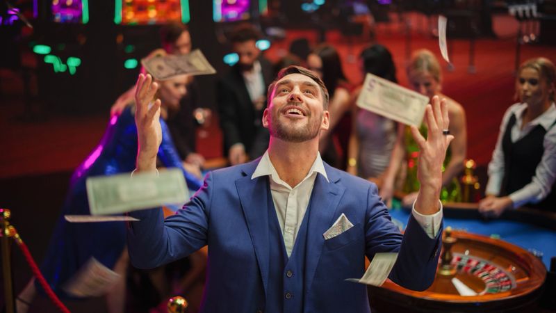 Happy Man in a Suit Standing in the Middle of a Modern Casino, Extending Arms, Welcoming a Money Rain.Young Male Smiling, Celebrating His Victory. Embodiment of a Successful Man