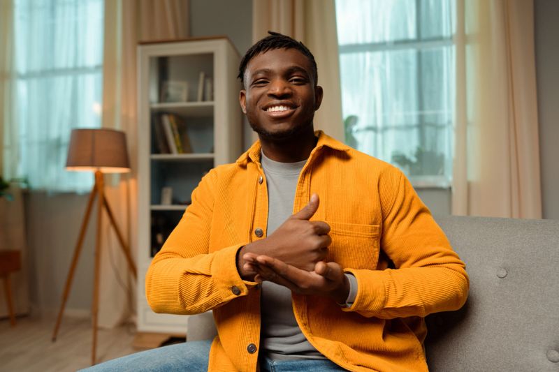 Portrait of smiling happy African American man speaking with sign language, showing finger symbols, wearing casual clothes, looking at camera while sitting at home. Communication concept