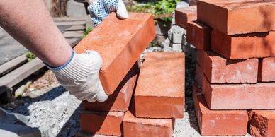 Hands in gloves stacking red bricks outdoors under sunlight.