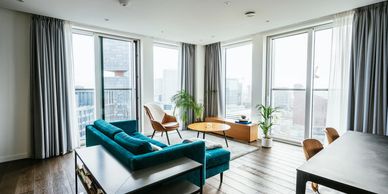 Modern living room with teal sofa, leather chair, and large windows overlooking cityscape.
