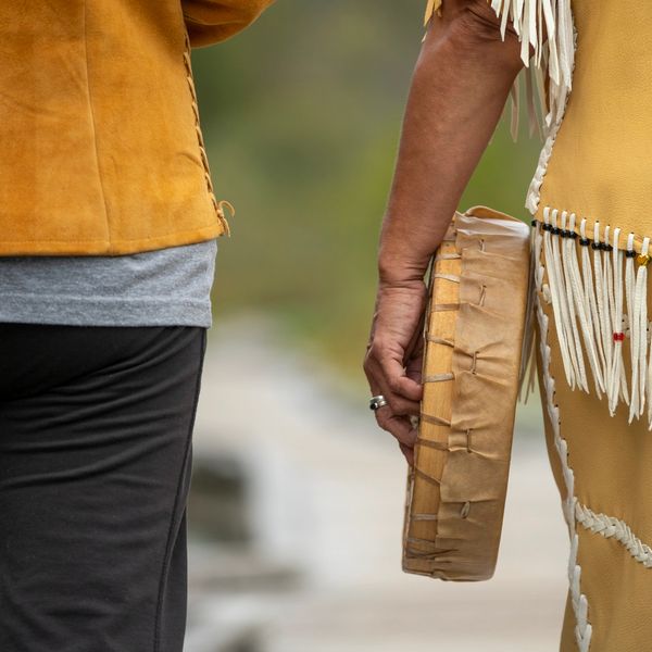 Two people walking, one holding a traditional drum and wearing fringed clothing.