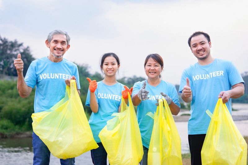 group of family volunteers in blue T-shirt standing,holding yellow plastic sorting bags and doing thumb up by the river