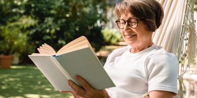 Elderly woman enjoying reading a book outdoors in a hammock.