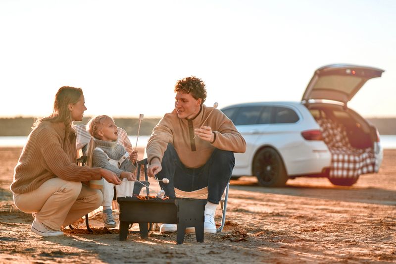 A family with a daughter sitting at the grill and roasting marshmallows. In the background is a car with an open trunk. Family traveling. Travel, vacation and tourism concept.