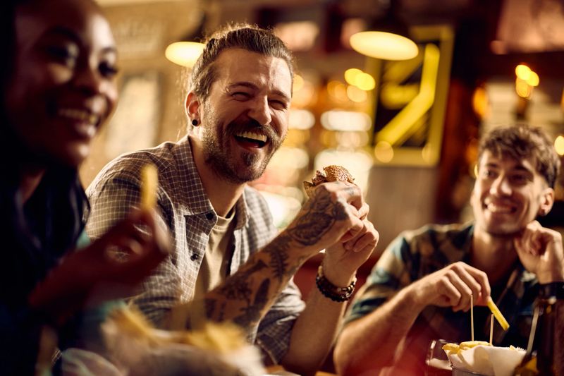 Cheerful man eating hamburger while gathering with friends in a bar.